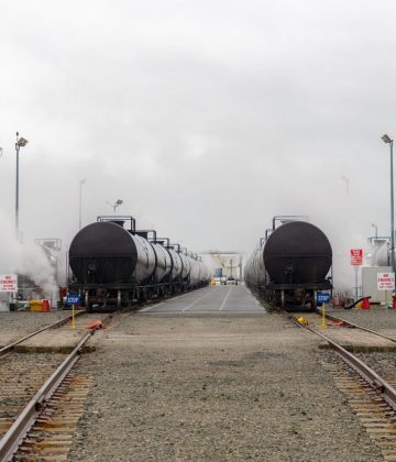 Railway tanker cars used to transport petroleum products. Several card visible. Fog or smoke rises from the cars. The cars are in the yard that loads them. Identifying marks removed. End view.